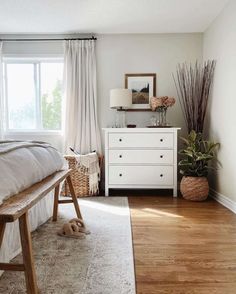 A woven basket with an indoor plant and tall ornamental stems stands beside a white dresser with black hardware. A clear glass table lamp and floral vase with pink hydrangeas decorate the top of the dresser. A wood-framed portrait hangs over the dresser along the pearl grey wall...   Image: ker_webb