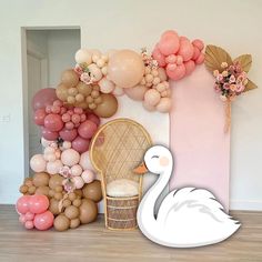 a white swan sitting on top of a wooden floor next to balloons and flowers in front of a pink wall