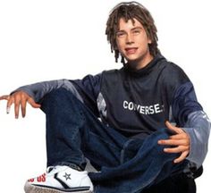 a man with dreadlocks sitting on top of a skateboard wearing a converse shirt