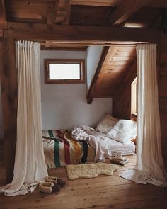 a bed sitting under a window in a bedroom next to a wooden floor covered in white curtains