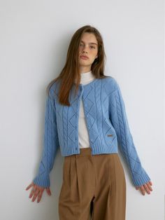 a woman standing in front of a white wall wearing a blue sweater and brown pants