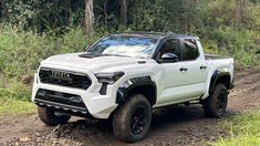 a white truck driving down a dirt road next to trees and bushes in the woods