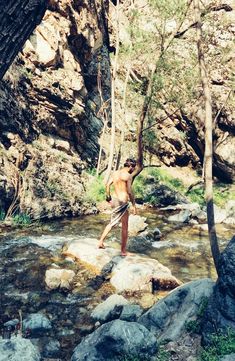 a shirtless man standing on top of a rock in a river surrounded by trees