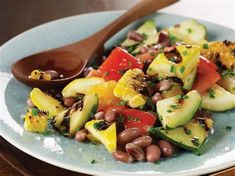 a salad with beans, zucchini and tomatoes on a blue plate next to a wooden spoon