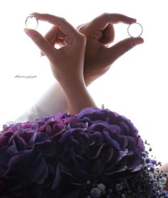 two hands holding rings over a bouquet of purple and white flowers in front of a window