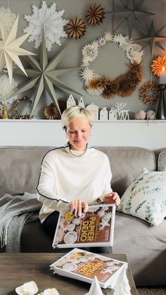 a woman sitting on a couch holding a magazine in front of a wall with paper snowflakes