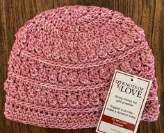 a pink crocheted hat sitting on top of a wooden table next to a tag