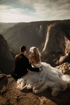 a bride and groom sitting on top of a mountain looking out at the valley below