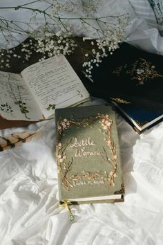 two books sitting on top of a bed next to flowers and an open book with the words little women written in it