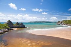 a sandy beach next to the ocean with rocks in the water and green hills on either side