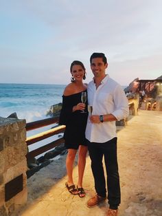 a man and woman standing next to each other near the ocean holding glasses of wine