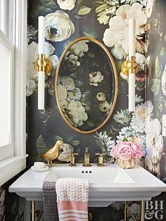 a white sink sitting under a mirror next to a wall covered in floral wallpaper