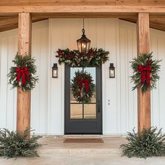 two christmas wreaths on the front door of a white house with red bows and lights