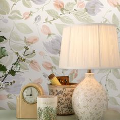 a table with a lamp, vase and other items on it in front of a floral wallpaper