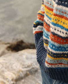 a person wearing a multicolored sweater standing next to a body of water and holding a cell phone