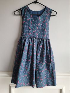 "-This adorable vintage dress is handmade and amazing. The fabric is likely cotton and is navy blue with a simple folk floral pattern in pinks and greens. It is sleeveless with buttons down the back with a sash at the back of waist. There are pockets on the front sides and buttons on the shoulders. Size is: 6x Length: 30.5\" + 1.5\" hem allowance Chest: 16\"" Cotton A-line Sundress For Garden Party, Cotton Floral Print Sleeveless Dress For Garden Party, Sleeveless Cotton Sundress With Floral Pattern, Blue Cotton Sleeveless Sundress, Blue Cotton Lined Sundress, Blue Floral Cotton Sundress, Cotton A-line Sleeveless Dress With Floral Print, Blue Floral Print Cotton Sundress, Sleeveless Fitted Cotton Floral Dress