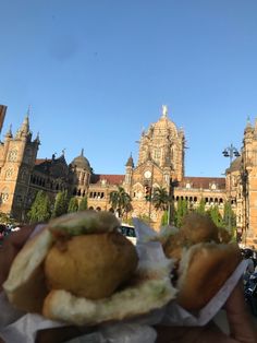 a hand holding a sandwich in front of a large building with towers and palm trees