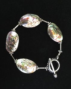 "This beautiful abalone shell bracelet is composed of gorgeous abalone flat ovals with silver spacers. The abalone beads are iridescent and you will see blue, pink, and green as light \"hits\" it. The shells on the bracelet are whole so they are the same on both sides. There are small silver tubes and silver spacers between each shell. The bracelet has a silver toggle closure. You could wear this interesting abalone sea shell bracelet with any outfit because it is neutral. This is a special brac Handmade Shell-shaped Silver Bracelets, Handmade Silver Shell Bracelets, Elegant Shell-shaped Bracelets, Silver Shell Bracelets Gift, Elegant Silver Abalone Shell, Handmade Silver Abalone Shell, Elegant Handmade Shell Bracelets, Elegant Iridescent Handmade Bracelets, Neutral Bracelet