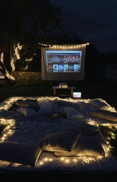 an outdoor movie screen is lit up at night with lights on the ground and pillows in front of it