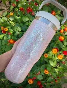 someone is holding a pink glittered water bottle in front of some plants and flowers