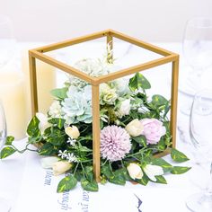 an arrangement of flowers and greenery in a glass box on a white table cloth