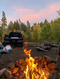 two people sitting around a campfire in the woods with a truck parked behind them