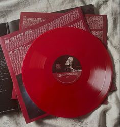 three red vinyl records sitting on top of a bed next to an open book and magazine