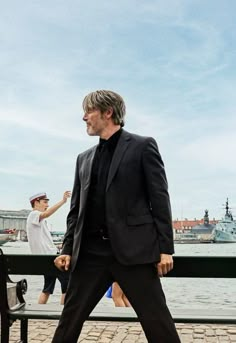 a man in a suit and tie walking by the water with his foot on a bench