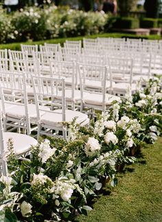 rows of white chairs lined up in the grass with flowers on each chair and greenery