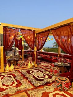 an outdoor living area with red curtains and rugs on the floor, overlooking the ocean