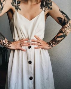 a woman with tattoos on her arms wearing a white dress and holding her hands behind her back