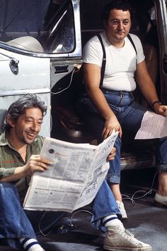 two men sitting in the back of a van reading newspapers and laughing at each other