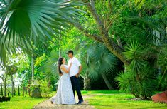 a man and woman are standing in the grass near some palm trees with their arms around each other