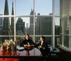 two people sitting at a table in front of large windows with cityscape behind them