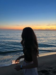 a woman standing on top of a beach next to the ocean holding a cell phone