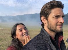 a man standing next to a woman on top of a lush green field covered in fog