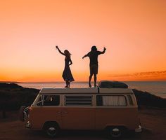 two people standing on top of an old vw camper van in front of the ocean