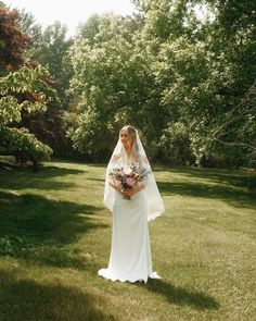 a woman in a wedding dress holding a bouquet and standing on the grass with trees behind her