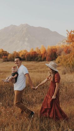 a man and woman holding hands while walking through a field with a baby in their arms