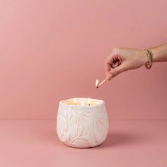 a person lighting a candle in a white ceramic bowl on a pink surface with one hand reaching for it