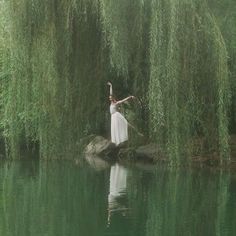 a woman in a white dress is standing on a rock by the water with willow branches