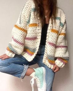 a woman sitting on top of a pair of blue jeans wearing white sneakers and a crocheted cardigan