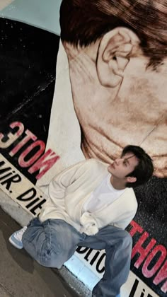 a young man sitting on the ground in front of a wall with a painting of a man's face