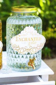 two glass jars with labels on them sitting on a white table in front of some bushes