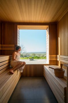 a woman sitting in a wooden sauna looking out the window at the landscape outside