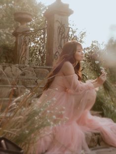 a woman in a pink dress sitting on the ground next to some tall grass and flowers