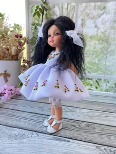 a doll with long black hair wearing a white dress and shoes on a wooden table