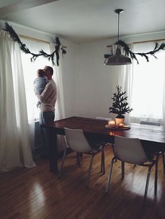 a man holding a baby standing in front of a dining room table with white chairs