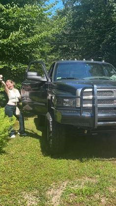 a woman standing next to a truck in the grass with her hand up and another person leaning on it