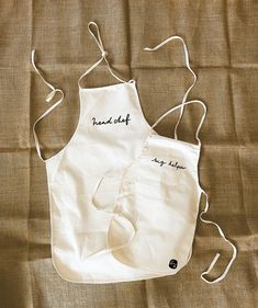 two white aprons sitting on top of a brown cloth covered tablecloth with writing